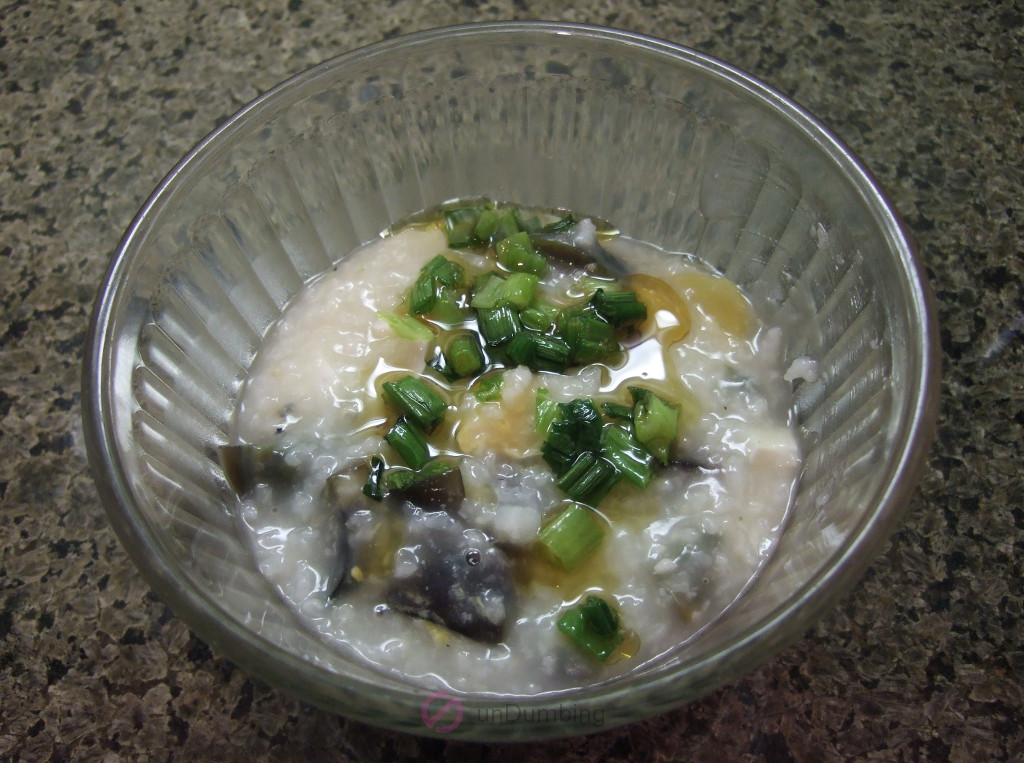 Glass bowl of porridge topped with thousand-year-old eggs, salted eggs, green onions, and sesame oil