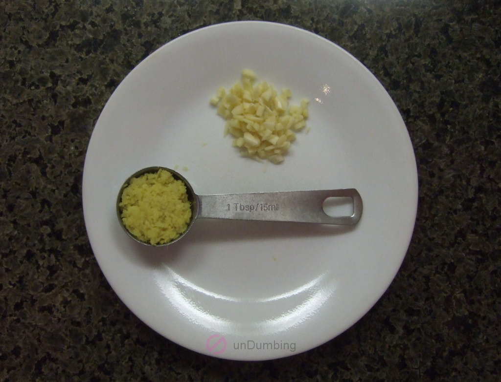 Minced garlic cloves and stainless steel measuring spoon of grated ginger on a white plate
